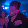 Un homme avec une barbe et les cheveux attachés en arrière, portant une chemise noire et une montre, allumant une cigarette avec un briquet.