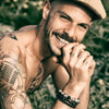 Un homme souriant avec une barbe, une moustache, des tatouages et des bracelets.