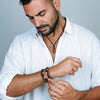 Un homme en chemise en lin blanc ajustant un bracelet en cuir sombre avec des accents en argent et en or sur son poignet gauche, portant également un collier en corde sombre avec un pendentif en pierre rouge rectangulaire.