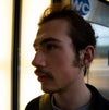 Un jeune homme aux cheveux foncés attachés en arrière, une moustache, et deux boucles d'oreilles en argent à son oreille gauche.