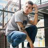 Un jeune homme avec des tatouages sur le bras, portant un t-shirt gris, un jean bleu et des baskets blanches.